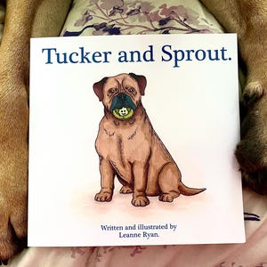 Op de afbeelding: Een kinderboek getiteld "Tucker and Sprout" met een illustratie van een bruine hond die een groene bal vasthoudt. De boekomslag heeft blauwe tekst op een witte achtergrond. De naam van de auteur staat ook op de omslag.