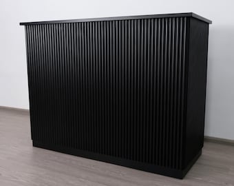 May include: A black cabinet with a vertical ribbed design. The cabinet has a flat top and a base, and it stands on a wooden floor. The overall design is modern and minimalist.