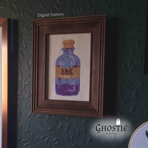 May include: Cross-stitch artwork of a potion bottle in a wooden frame. The bottle is blue with a purple base and a cork stopper. The label reads "FAE". The words "Digital Pattern" are above the frame.