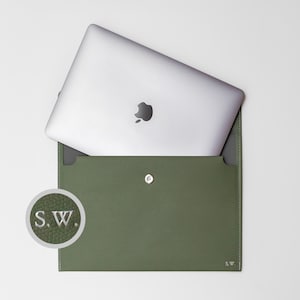 Könnte beinhalten: Eine grüne Ledertasche für Laptops mit Druckknopfverschluss und den eingeprägten Initialen "S.W." auf der Vorderseite.