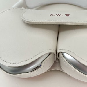 May include: White leather case with two compartments for wireless earbuds. The case has a flap with the initials "S.W." and a pink heart.