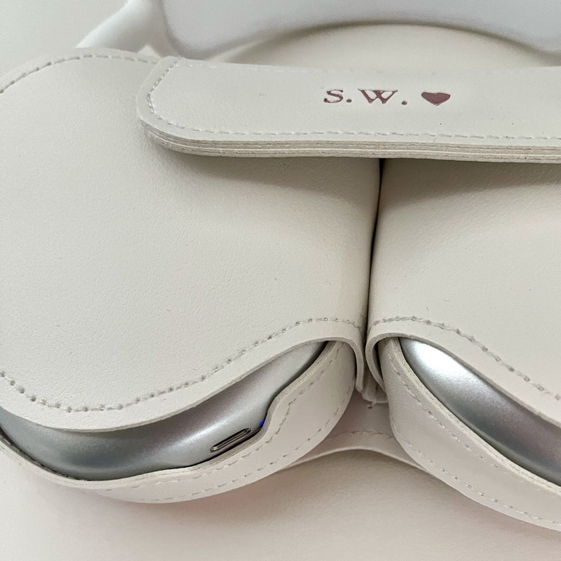May include: White leather case with two compartments for wireless earbuds. The case has a flap with the initials "S.W." and a pink heart.