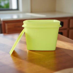 May include: A square, lime green plastic container with a matching lid. The lid is partially open, revealing the interior of the container. The container is sitting on a wooden surface, with a blurred background of a kitchen.