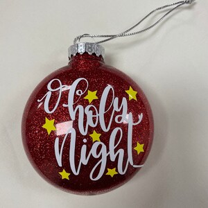 Könnte beinhalten: Roter Glitzer-Weihnachtsbaumschmuck mit den Worten "O Holy Night" in weißer Schrift und gelben Sternen.