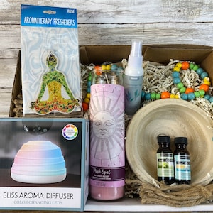 May include: A gift box filled with aromatherapy items, including a diffuser, essential oils, a candle, and a beaded necklace. The box is filled with shredded paper and has a wooden lid.