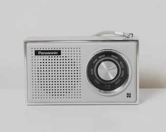Może przedstawiać: Srebrne radio Panasonic z kratką głośnika, pokrętłem strojenia i chowaną anteną. Nazwa marki jest wydrukowana z przodu. Radio stoi na białej powierzchni na białym tle.