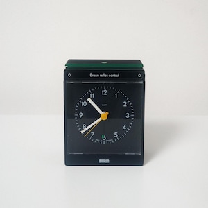 Może przedstawiać: Czarny i zielony zegar alarmowy Braun reflex control z białą tarczą i żółtymi wskazówkami. Zegar ma wyświetlacz cyfrowy, a na dole nadrukowany jest napis "Braun".