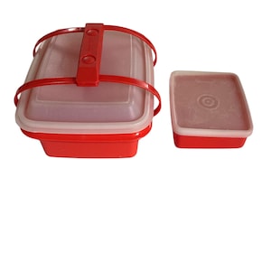 May include: Red and clear plastic food storage containers. The larger container has a red handle and lid. The smaller container has a red base and a clear lid. Both containers are rectangular.