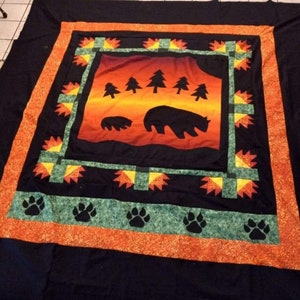 May include: A black and orange quilt featuring a design of bears and trees against a sunset backdrop. The quilt has a border of green and orange with paw prints.
