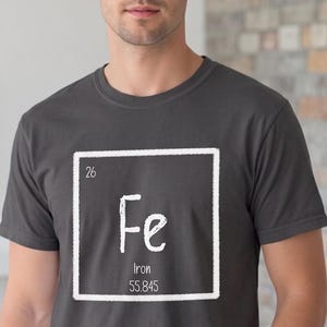 May include: Dark grey t-shirt featuring the periodic table symbol for iron, Fe, in a white square. The shirt also displays the atomic number 26 and the atomic mass 55.845. The person is wearing blue jeans.