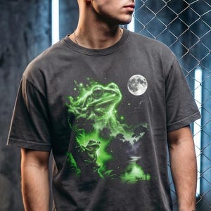 May include: Dark gray t-shirt featuring a vibrant green frog graphic with a full moon. The design includes a detailed illustration of a frog with a celestial background. The shirt is a casual wear item.
