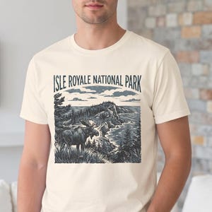 May include: Cream-colored t-shirt featuring a detailed illustration of Isle Royale National Park. The graphic depicts a moose near a shoreline with trees and the text "ISLE ROYALE NATIONAL PARK" above the image. The shirt is worn by a person.