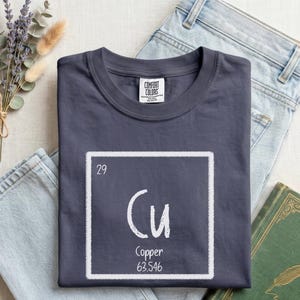 May include: A denim blue t-shirt with a white square containing the element symbol "Cu" for copper. The atomic number 29 and atomic mass are also displayed. The word "Denim" is printed at the bottom of the shirt. The shirt is laid on top of a pair of light blue jeans.