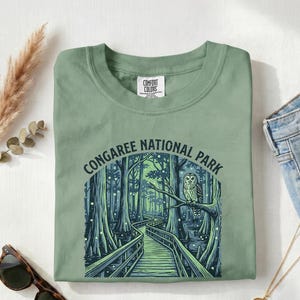 May include: Light green t-shirt with a graphic of Congaree National Park, featuring a boardwalk through a swamp and an owl perched on a branch. The text "CONGAREE NATIONAL PARK" is arched above the image. The words "Light Green" are printed below the graphic.