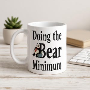 May include: White ceramic mug with the text "Doing the Bear Minimum" in black. A cartoon panda wearing a red tie and holding a briefcase is featured on the mug. The mug has a curved handle.