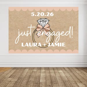 May include: A large sign with the words "just engaged!" in a cursive font, along with the names "LAURA + JAMIE" in a block font. The sign features a diamond ring graphic and the date "5.20.26". The background is a light brown color with a scalloped border.