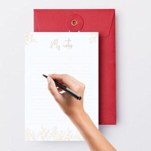 May include: A white notepad with the words "My notes" in gold script, with gold floral accents, is held by a person writing with a black pen. A red envelope is in the background.