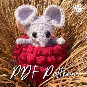 May include: A crocheted mouse in a red heart-shaped basket. The mouse is gray with black eyes and a pink nose. The basket is made of red yarn and has a textured surface. The text "PDF Pattern" is written in white on the bottom of the image.