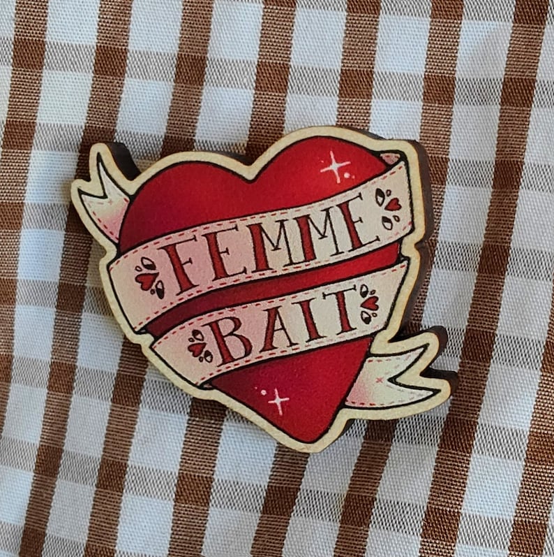 May include: A red heart-shaped wooden pin with a white ribbon wrapped around it. The ribbon has the words "FEMME BAIT" written on it in black.