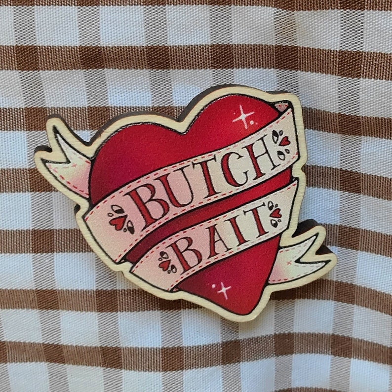 May include: A wooden heart-shaped pin with a red heart and a white banner that says "Butch Bait".
