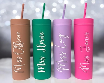 May include: Four personalized tumblers with straws. The tumblers are in different colors: brown, green, purple, and pink. Each tumbler has a name written on it in white lettering: Miss Officer, Mrs Horne, Miss Lucy, and Mrs Jones.