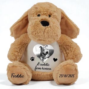 May include: A soft, tan-coloured dog toy with large ears and a black nose. It features a white heart with a black and white dog photo, paw prints, and a heart. Text includes "A cuddle from heaven", "Freddie", and "23/10/2025".