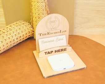 May include: A wooden stand with the text "The Etched Lab" and "Business Card" on it. The stand has a slot for a business card and a space for a white square device with the text "TAP HERE" below it. The stand is on a brown leather surface.