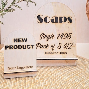 May include: Two wooden signs with rounded tops. The larger sign says "Soaps" in black lettering and has the text "Single £4.95 Pack of 3 £12- Bubbles Wishes". The smaller sign says "NEW PRODUCT" in black lettering and has the text "Your Logo Here".