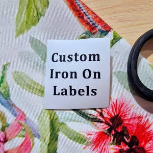 May include: A square white label with the black text Custom Iron On Labels printed in a serif font. The label is placed on a white fabric featuring a colourful botanical print of green leaves and vibrant red flowers, with a black circular object nearby.
