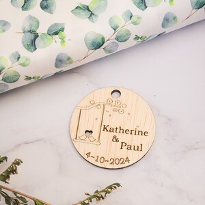 May include: A round wooden tag with a laser-engraved design of a swing set with two birds and a heart. The text "Katherine & Paul 4-10-2024" is engraved on the tag.