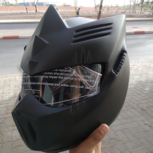 Può includere: Casco da moto nero con un design a forma di gatto e una visiera trasparente. Il casco ha una finitura opaca e caratteristiche angolari e affilate.