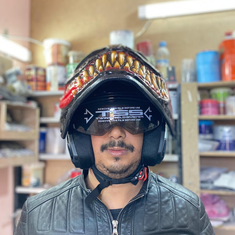 May include: A black motorcycle helmet with a red and yellow monster design. The helmet has a clear visor with the text "TSS Twin Shield System" and "Remove this film before use" printed on it.