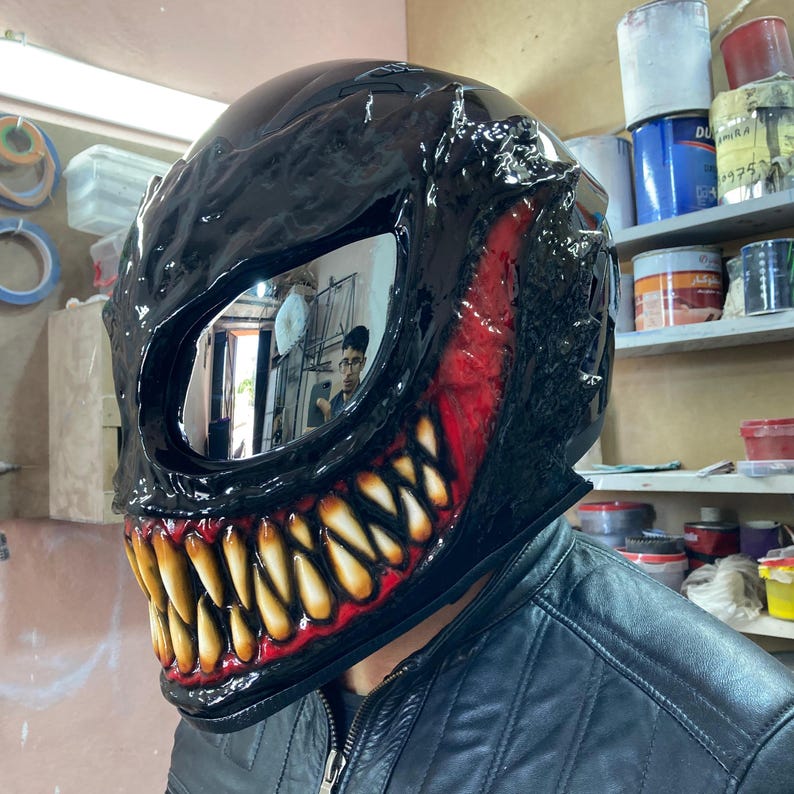 May include: A black motorcycle helmet with a red and yellow Venom design. The helmet has a mirrored visor and a large, toothy grin.