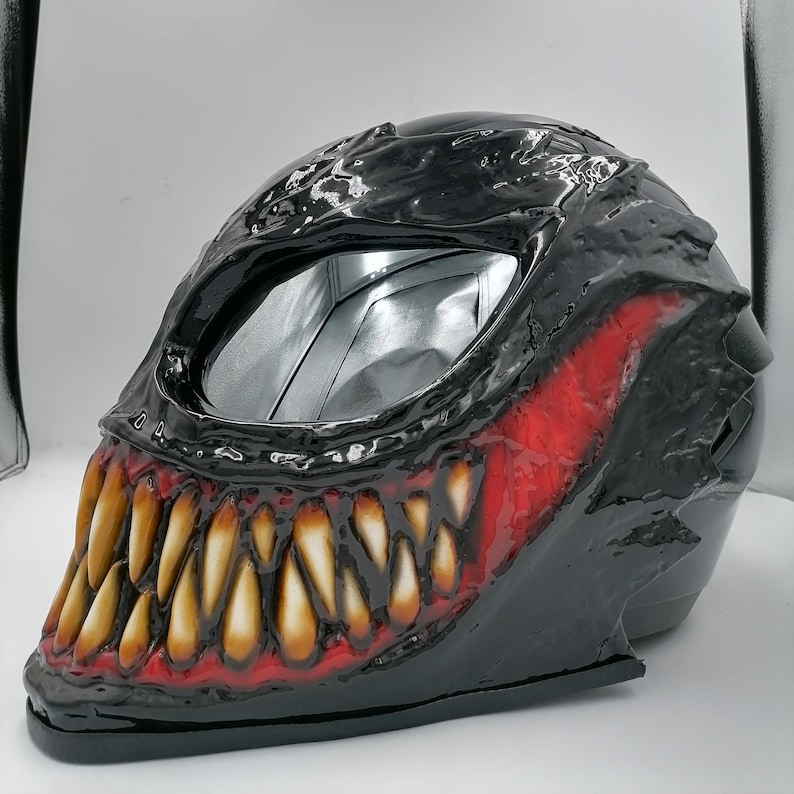 May include: Black motorcycle helmet with a red and yellow Venom design. The helmet has a large, toothy grin with sharp teeth.