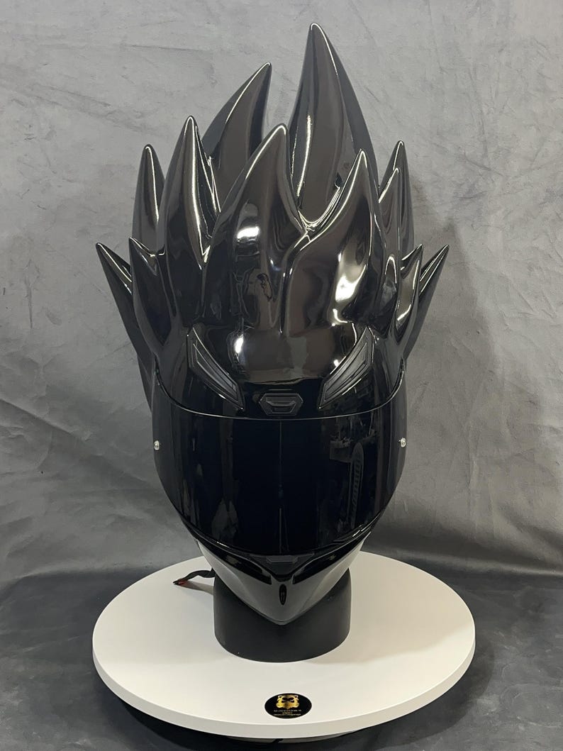 Op de afbeelding: Een glanzend zwarte motorhelm met een stekelig haarontwerp. De helm heeft een donker vizier en wordt gepresenteerd op een witte, ronde standaard. Het ontwerp is ge&iuml;nspireerd op een personage uit een animatieserie.