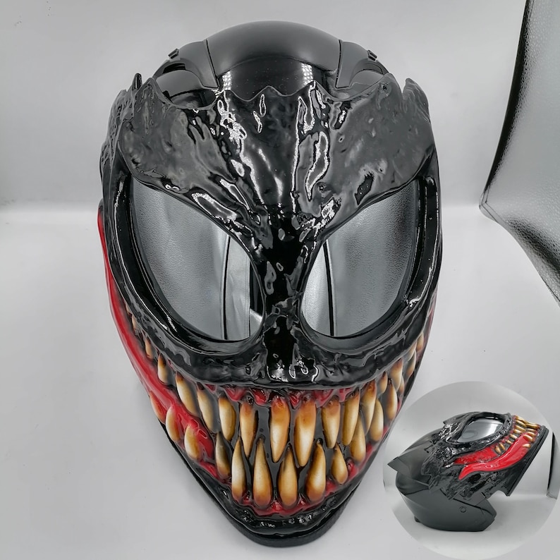 May include: Black motorcycle helmet with a Venom design. The helmet has a black and red paint scheme with a detailed Venom face and sharp teeth.