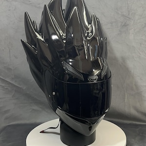 Op de afbeelding: Een glanzend zwarte motorhelm met een uniek, stekelig ontwerp aan de bovenkant, dat lijkt op gestileerd haar. De helm heeft een donker vizier en wordt gepresenteerd op een witte en zwarte standaard. De algehele esthetiek is futuristisch en edgy.