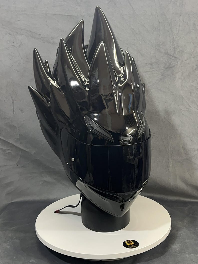 Op de afbeelding: Een glanzend zwarte motorhelm met een uniek, stekelig ontwerp aan de bovenkant, dat lijkt op gestileerd haar. De helm heeft een donker vizier en wordt gepresenteerd op een witte en zwarte standaard. De algehele esthetiek is futuristisch en edgy.