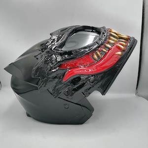May include: Black motorcycle helmet with a red and yellow Venom design. The helmet has a clear visor and a black chin strap.