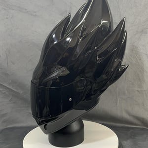 Op de afbeelding: Een glanzend zwarte motorhelm met een uniek, stekelig ontwerp. De helm heeft een donker vizier en wordt gepresenteerd op een witte, cirkelvormige standaard. De algehele esthetiek is futuristisch en edgy.