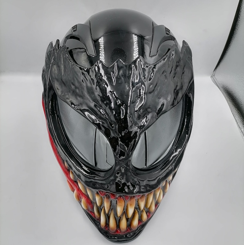 May include: A black motorcycle helmet with a Venom-inspired design. The helmet features a black and red paint scheme with a detailed Venom face mask.