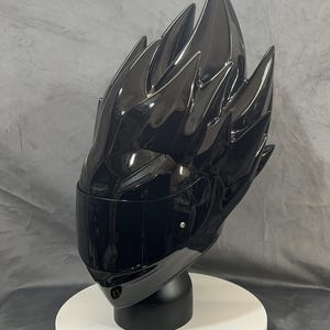 Op de afbeelding: Een glanzend zwarte motorhelm met een uniek, stekelig ontwerp dat lijkt op gestileerd haar. De helm heeft een getint vizier en wordt gepresenteerd op een witte, cirkelvormige basis met een zwarte standaard. De algehele esthetiek is futuristisch en gedurfd.