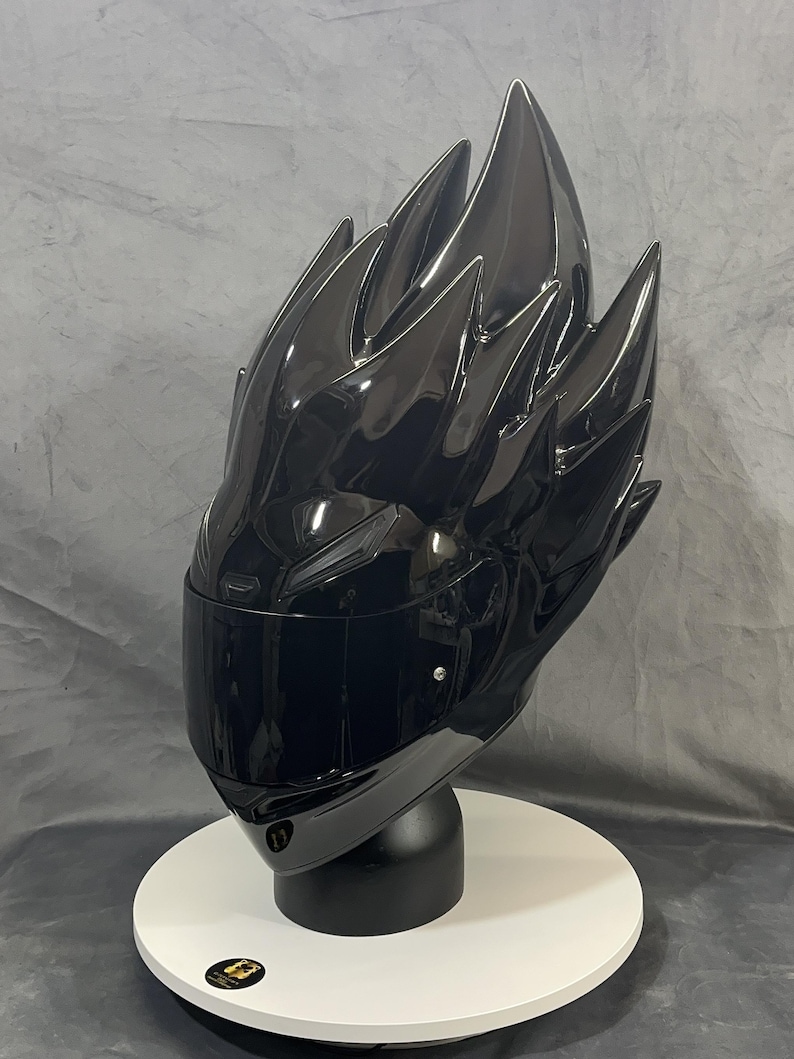 Op de afbeelding: Een glanzend zwarte motorhelm met een uniek, stekelig ontwerp dat lijkt op gestileerd haar. De helm heeft een getint vizier en wordt gepresenteerd op een witte, cirkelvormige basis met een zwarte standaard. De algehele esthetiek is futuristisch en gedurfd.