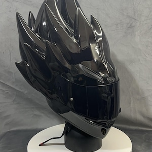 Op de afbeelding: Een glanzend zwarte motorhelm met een uniek, stekelig ontwerp dat lijkt op gestileerd haar. De helm heeft een donker vizier en wordt gepresenteerd op een witte, cirkelvormige standaard. De algehele esthetiek is futuristisch en gedurfd.