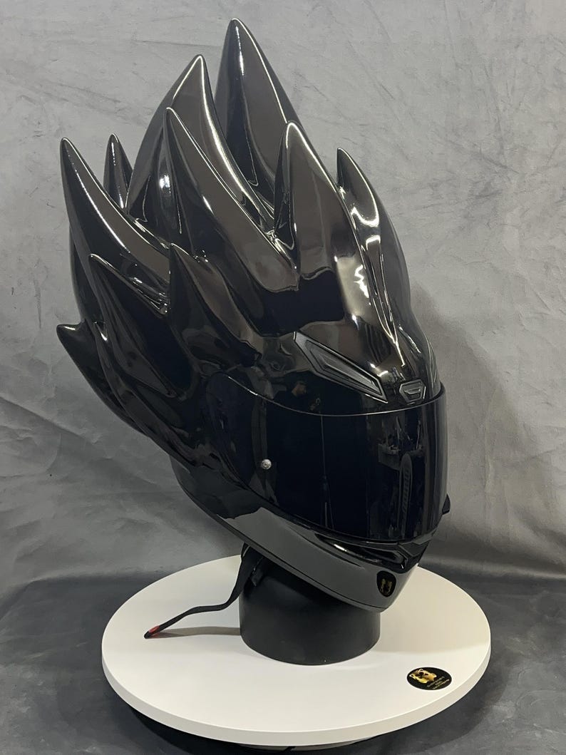 Op de afbeelding: Een glanzend zwarte motorhelm met een uniek, stekelig ontwerp dat lijkt op gestileerd haar. De helm heeft een donker vizier en wordt gepresenteerd op een witte, cirkelvormige standaard. De algehele esthetiek is futuristisch en gedurfd.