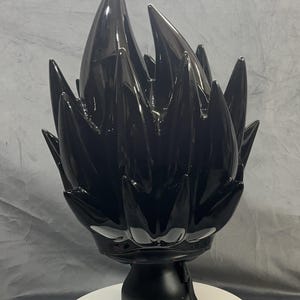 Op de afbeelding: Zwarte, stekelige sculptuur die gestileerd haar voorstelt, gemonteerd op een zwarte basis en een wit, cirkelvormig platform. De sculptuur heeft een glanzende afwerking en een riem met een rode accent.