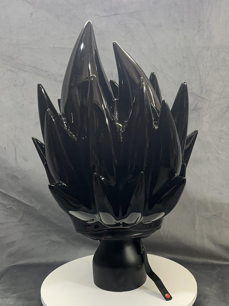 Op de afbeelding: Zwarte, stekelige sculptuur die gestileerd haar voorstelt, gemonteerd op een zwarte basis en een wit, cirkelvormig platform. De sculptuur heeft een glanzende afwerking en een riem met een rode accent.