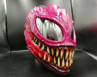 May include: A magenta-colored, monster-themed helmet with large, white teeth and a red mouth. The helmet has large eye openings with a reflective silver material inside. The overall design is textured and detailed, giving it a menacing appearance.