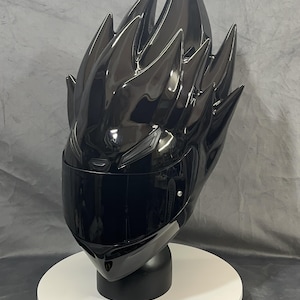 Op de afbeelding: Een glanzende zwarte motorhelm met een uniek, stekelig ontwerp dat lijkt op gestileerd haar. De helm heeft een donker vizier en wordt gepresenteerd op een witte, cirkelvormige standaard. De algehele esthetiek is futuristisch en opvallend.