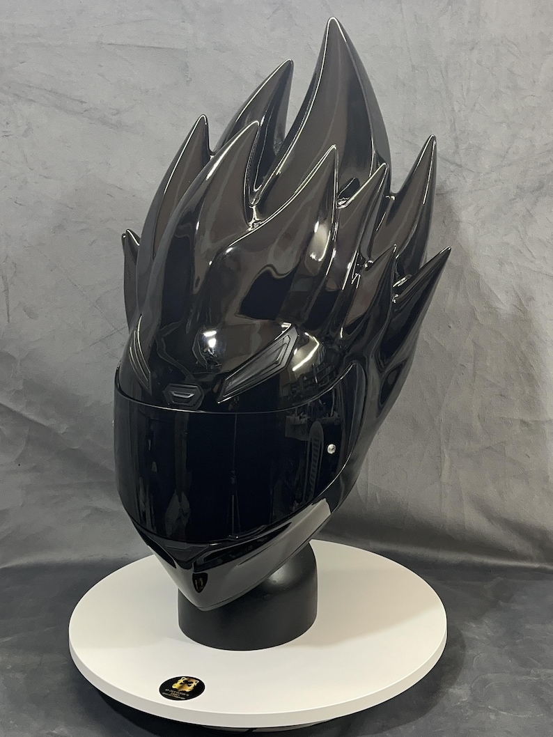 Op de afbeelding: Een glanzende zwarte motorhelm met een uniek, stekelig ontwerp dat lijkt op gestileerd haar. De helm heeft een donker vizier en wordt gepresenteerd op een witte, cirkelvormige standaard. De algehele esthetiek is futuristisch en opvallend.